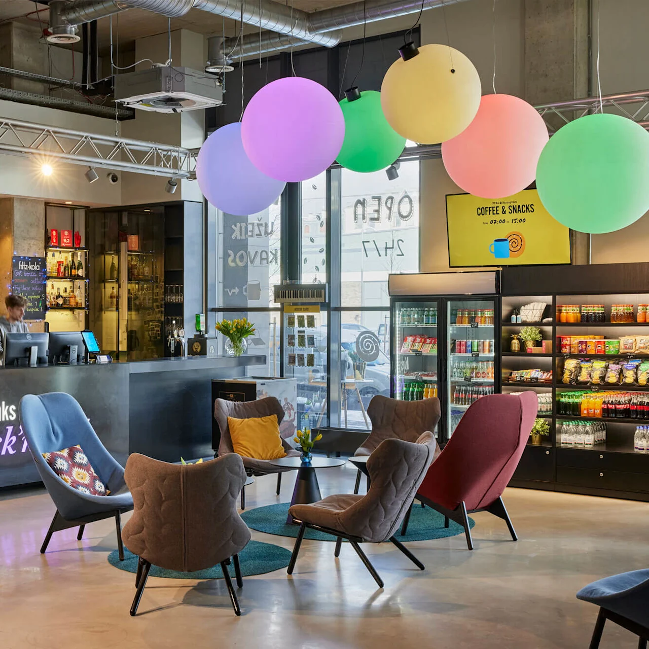 Reception med två anställda som hjälper gäster, omgiven av färgglada stolar och ballonger. I närheten finns hyllor med snacks och drycker. Synlig text: “Keys, drinks Rock’n”, “COFFEE & SNACKS 09.00 - 19.00”.