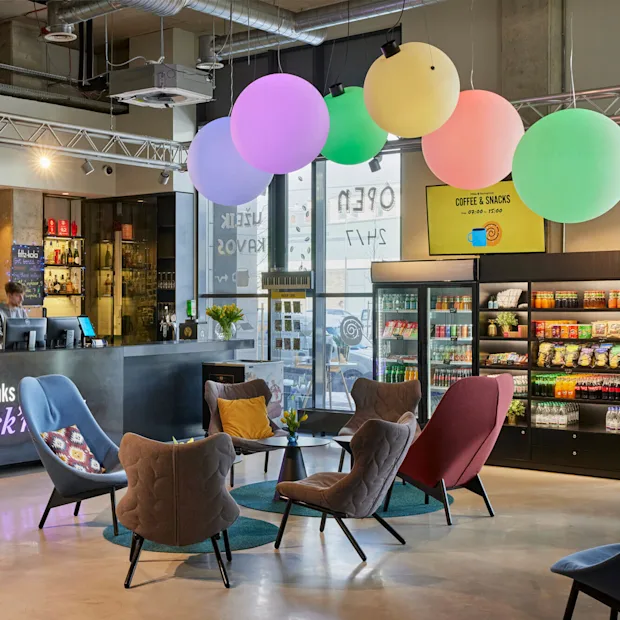 Reception desk with two attendants assisting guests, surrounded by colorful chairs and balloons. Nearby, shelves display snacks and drinks. Visible text: “Keys, drinks Rock’n”, “COFFEE & SNACKS 09.00 - 19.00”.