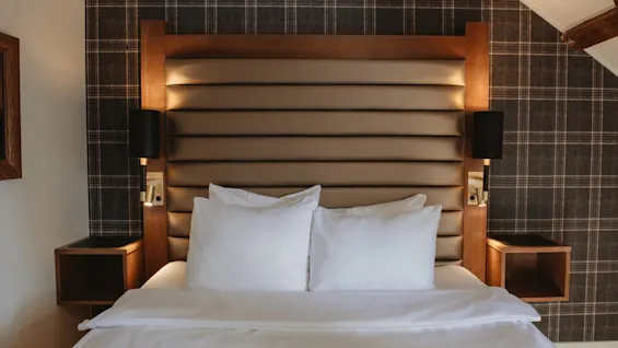 Et hyggeligt hotelværelse med en seng med hvidt sengetøj, en polstret sengegavl og ternet tapet, der indbyder til et afslappende ophold.