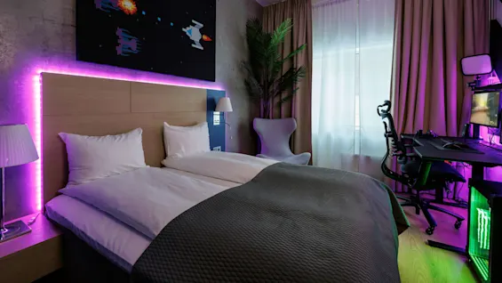 A bed with white sheets is illuminated by purple LED lights beneath a space-themed picture. Nearby, a gaming setup with a chair and computer is visible, framed by curtains.