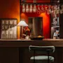 En køkkenbar med en grøn stol, belyst af en lampe, har hængende glas og et "HOME SWEET HOME" skilt. En person ses i baggrunden.