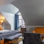 Et koselig soverom med en stor seng, polstret hodegjerde og fargerike puter ved siden av et vindu med blå gardiner. Ved siden av finnes en oransje sofa og et tre skrivebord.