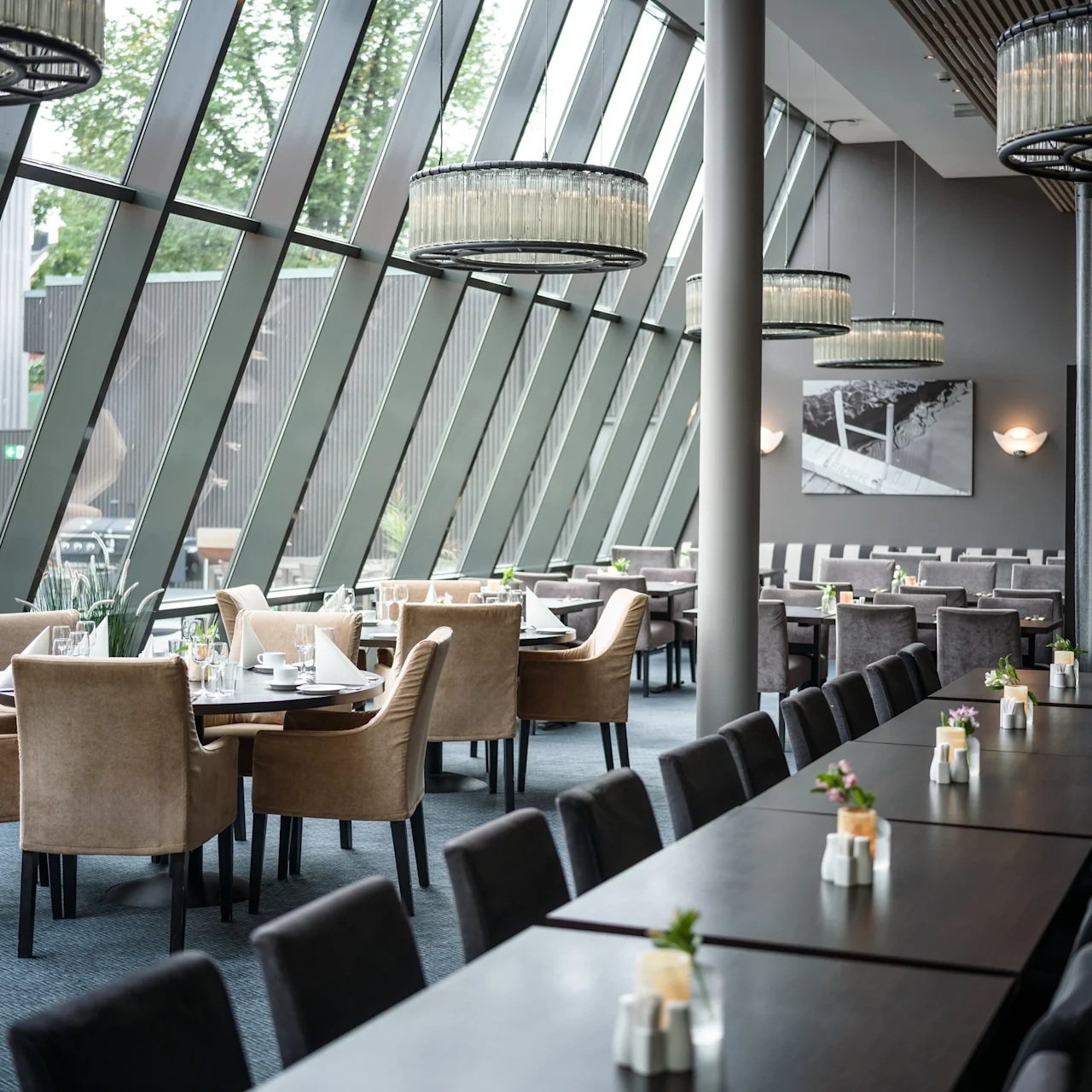 Eleganta matsalsstolar omger bord i en modern restaurang. Ljuskronor hänger ovanifrån, och lutande fönster visar frodig grönska utanför.
