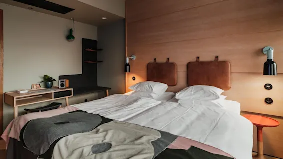 A neatly made double bed with white sheets, leather headboards, and modern lamps in a minimalist hotel room, featuring a wooden desk with a small plant and books.