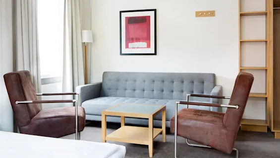 En grå sofa står mellom to brune lenestoler rundt et trebord i et lyst rom med et vindu. Et rødt abstrakt maleri pryder veggen.
