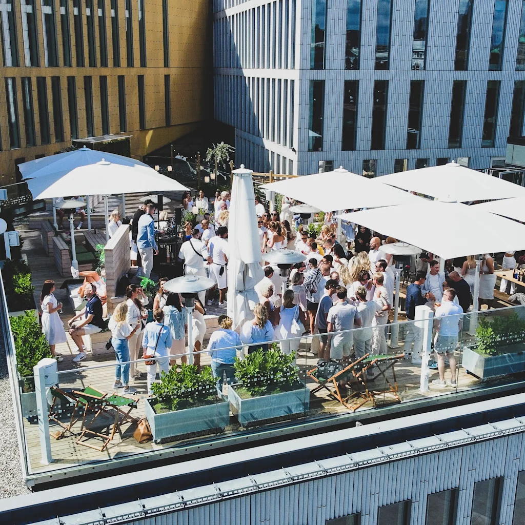 Ett livligt takterrassevent med många människor som umgås under vita parasoller och tak, omgivna av moderna byggnader.