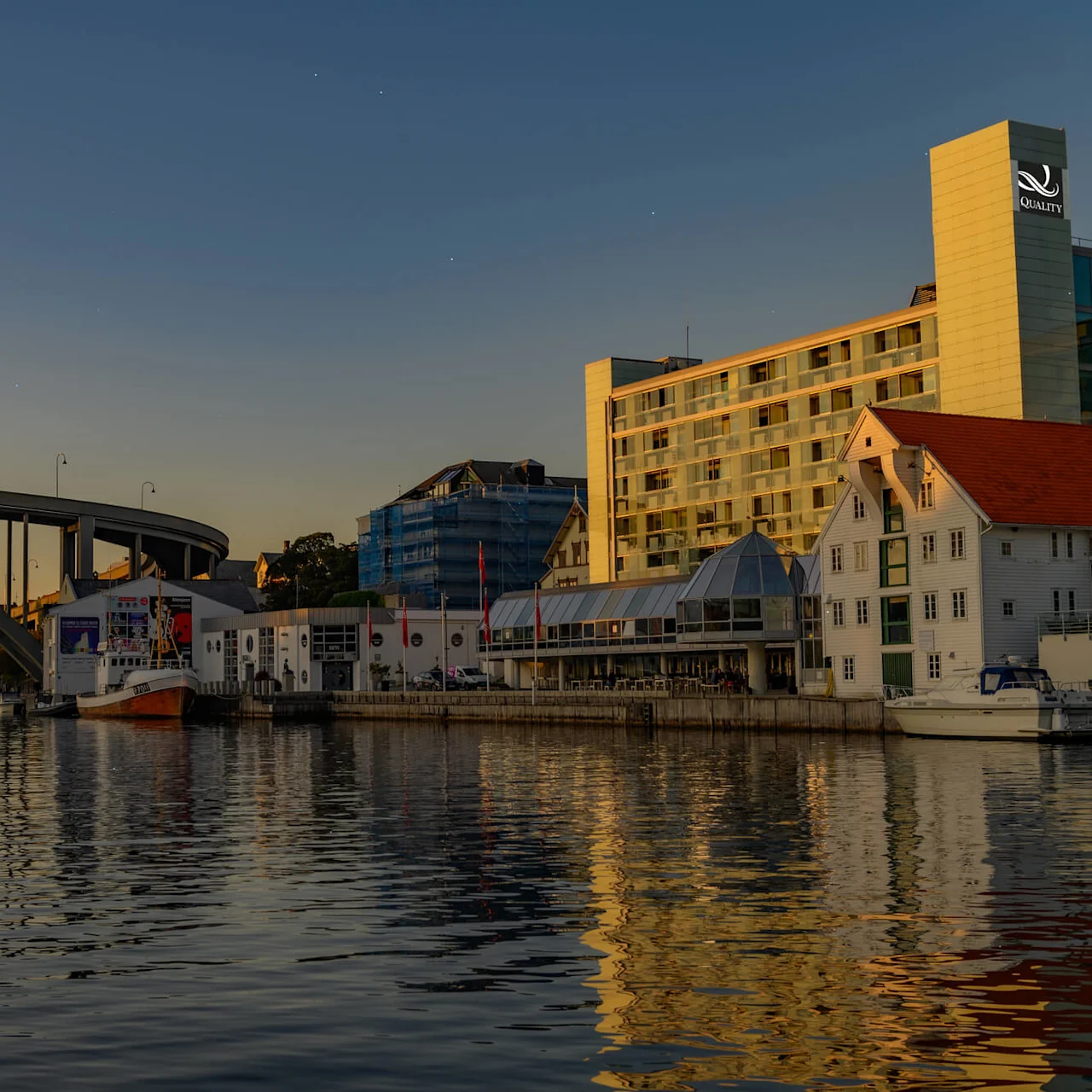 En stor hotellbyggnad med "QUALITY"-skylt blickar ut över ett område vid vattnet vid solnedgång. Båtar är förtöjda nära strandlinjen, och en välvd bro sträcker sig över vattnet.