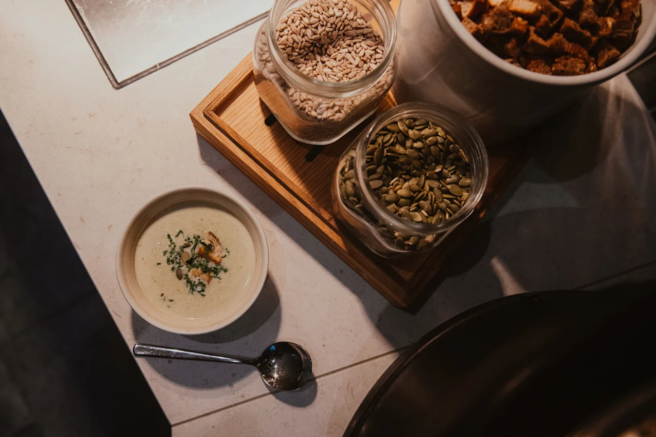 En krämig soppa i en skål med en sked, bredvid burkar med frön och krutonger, redo för en utsökt måltid.