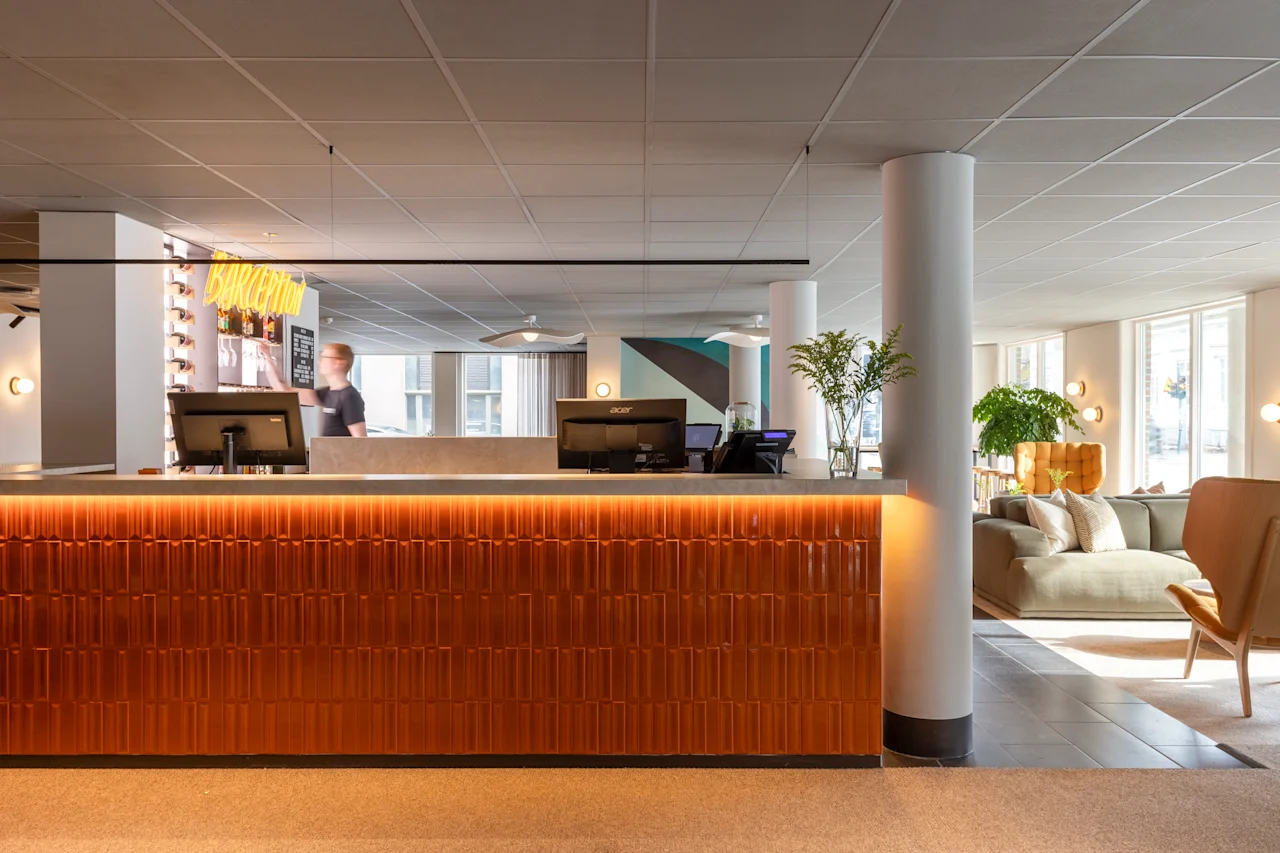 Reception med datorer och en person bakom. Neonljusskylt lyder "BOKA RUM." Modern lounge med soffor och växter. Stora fönster släpper in naturligt ljus.