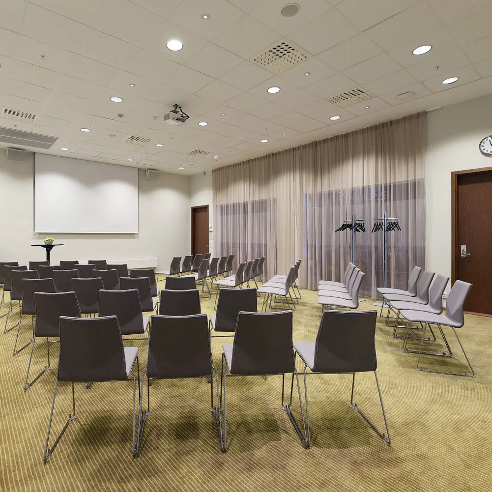 Chairs arranged in rows face a blank projector screen in a classroom setting with beige curtains, a wall clock, and a cabinet with a clock at 11:00.