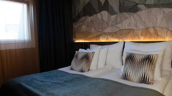 Et komfortabelt hotellrom med seng, turkis dynetrekk, dekorative puter og fjellmaleri med varm bakgrunnsbelysning.