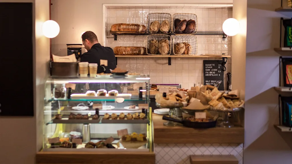 A cafe counter displays pastries and bread under glass. A person works behind the counter. Shelves with bread loaves are in the background. Warm lighting enhances the cozy atmosphere.