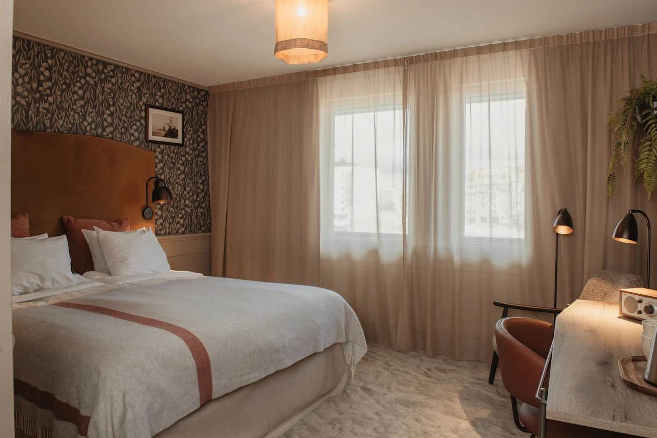 Et hyggeligt hotelværelse med en stor seng, et skrivebordsområde og lyse vinduer med lette gardiner, der giver en følelse af komfort og afslapning.