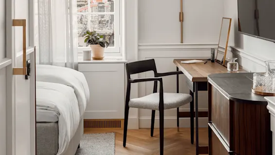 A cozy bedroom features a neatly made bed, a wooden desk with a chair, and a window with plants on the sill. Warm lighting enhances the modern, minimalistic decor.