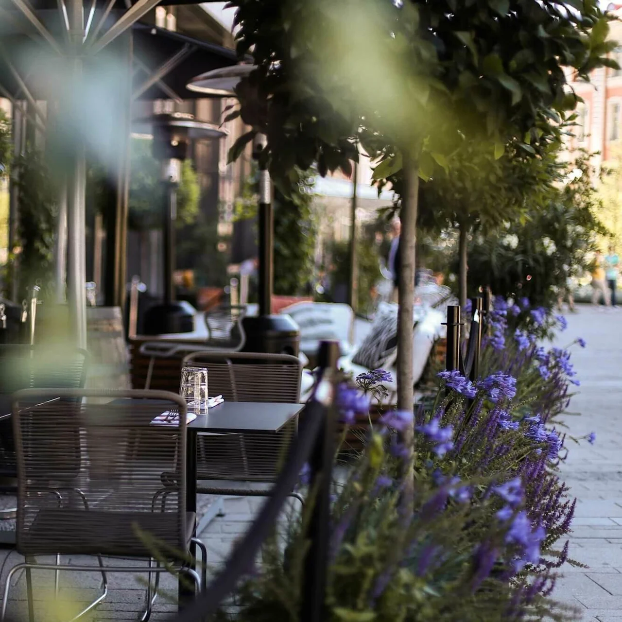 Uterestaurantbord med stoler plassert langs en gangsti. Lilla blomster og grønt rammer inn scenen, noe som skaper en frodig, innbydende atmosfære på en solrik dag.