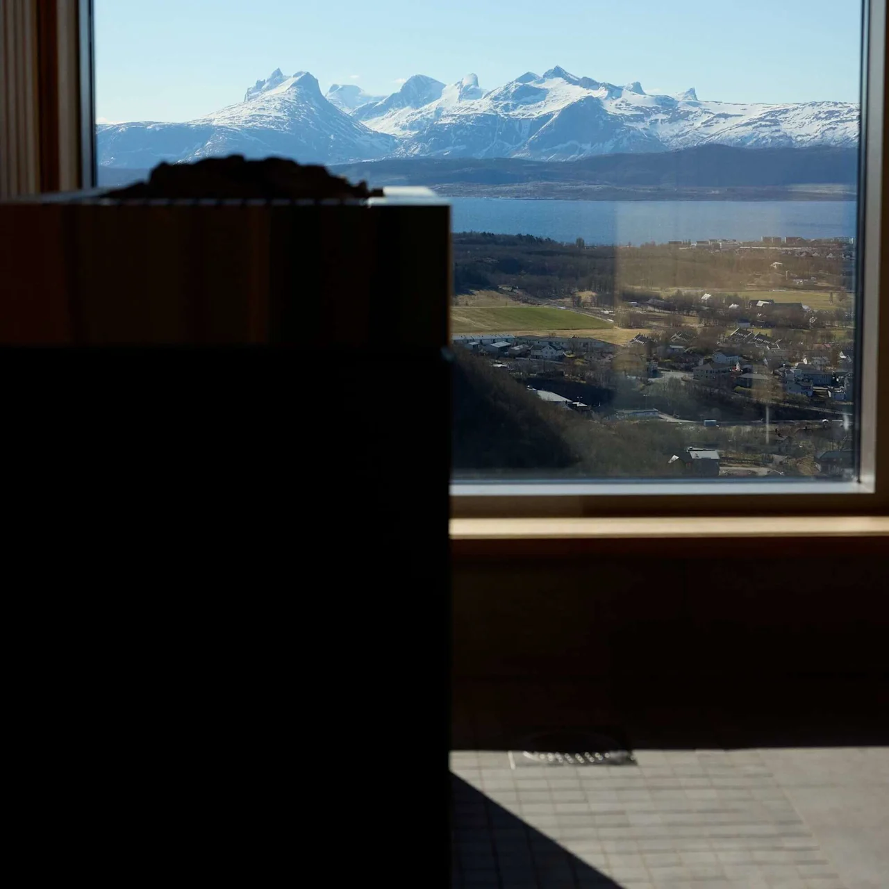 A sauna stove is silhouetted in the foreground, with snow-capped mountains visible through a large window, overlooking a town by a clear blue sky.