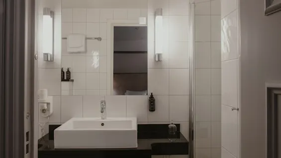 Et rent og moderne hotelbadeværelse med hvid vask, stort spejl og flisebelagte vægge, med toiletartikler og bruser.
