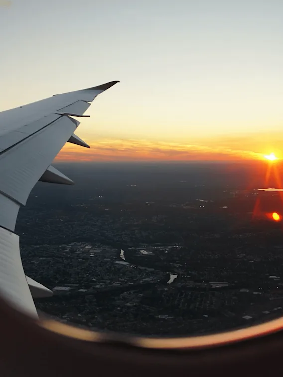 Flygplansvinge sträcker sig utåt, svävande ovanför en utbredd stadsvy. En livlig solnedgång färgar himlen med orange toner, med reflektioner på ett avlägset vattendrag.