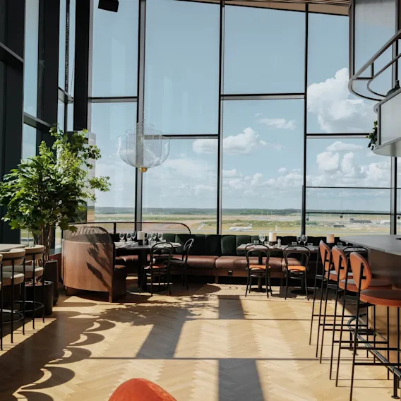 Et romslig moderne barområde med høye stoler og bord, omgitt av store glassvinduer som avslører en flyplassbane under en blå himmel med spredte skyer.