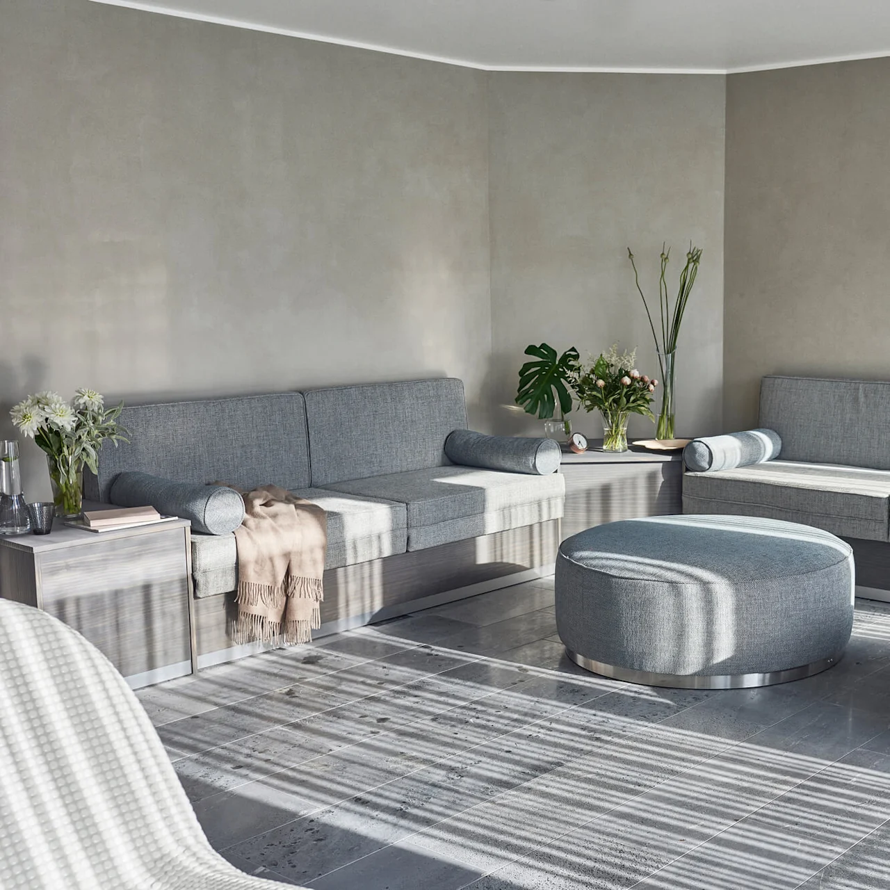 A modern spa reception with a large gray sectional sofa, two round ottomans, and decorative plants on a patterned rug and tiled floor.