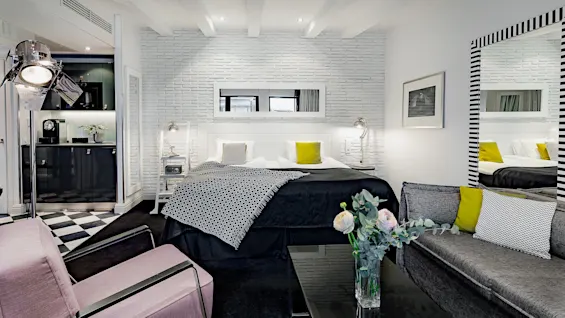 Et moderne soverom har en dobbeltseng med geometriske puter, omgitt av stilige møbler og myk belysning. Et kjøkkenområde og en stilig sofa med blomster gir en koselig atmosfære.