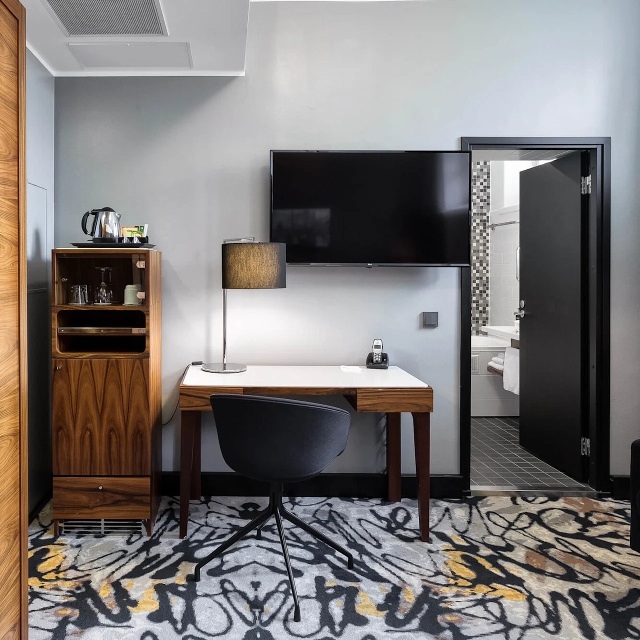 A modern hotel room with a desk, chair, and flat-screen TV; a minibar cabinet and open bathroom door reveal a neat, tiled interior.