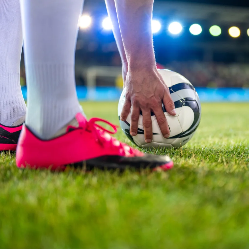 A person placing their hand on a soccer ball, wearing pink cleats on grass with stadium lights in the background.