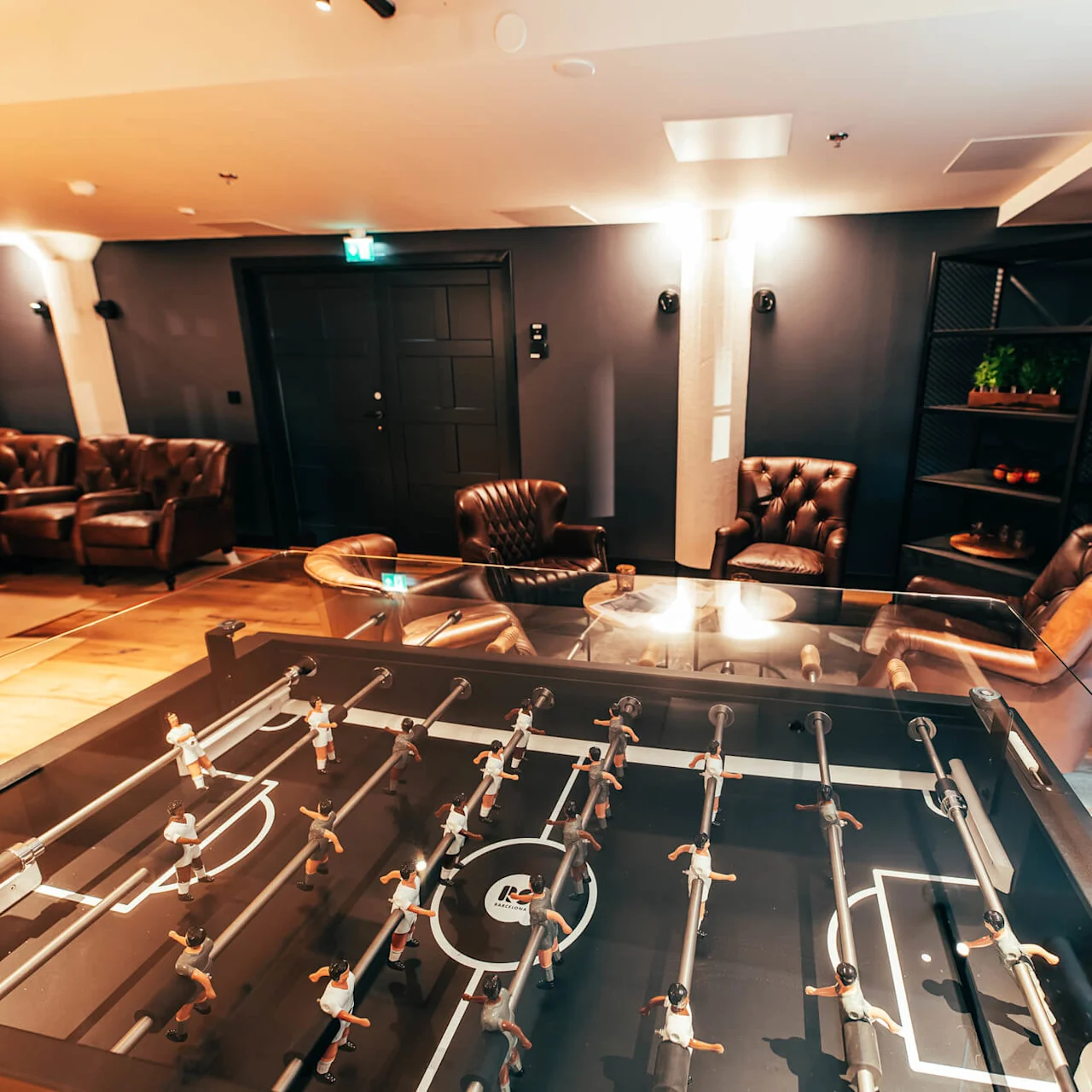Fotballspillbord står i sentrum, omgitt av brune lærstoler og en svart hylle med planter, i en svakt opplyst lounge med tregulv og moderne atmosfære.
