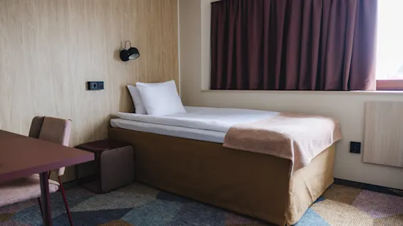 Single bed with white sheets and brown blanket sits against a wooden wall; desk and chair nearby. A dark curtain covers the window, providing a cozy ambiance.