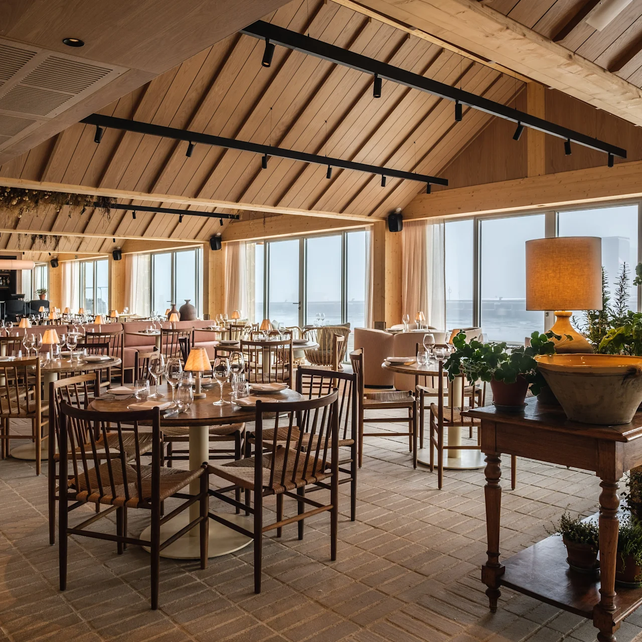A large dining room with wooden tables and chairs is arranged neatly. Large windows provide a view of the ocean. Potted plants decorate a side table, creating a cozy ambiance.