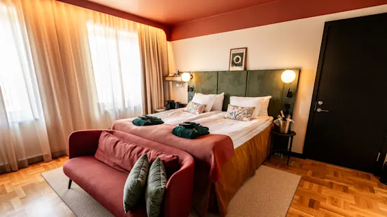 A double bed with floral pillows rests against a green headboard, beside a red sofa with green pillows; warm lighting fills the room, enhancing wooden flooring.