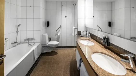 Badrum med brun bänkskiva med två handfat, intill en vit kaklad vägg. Ett badkar och toalett är placerade i närheten, med en spegel ovanför handfaten som reflekterar rummet.