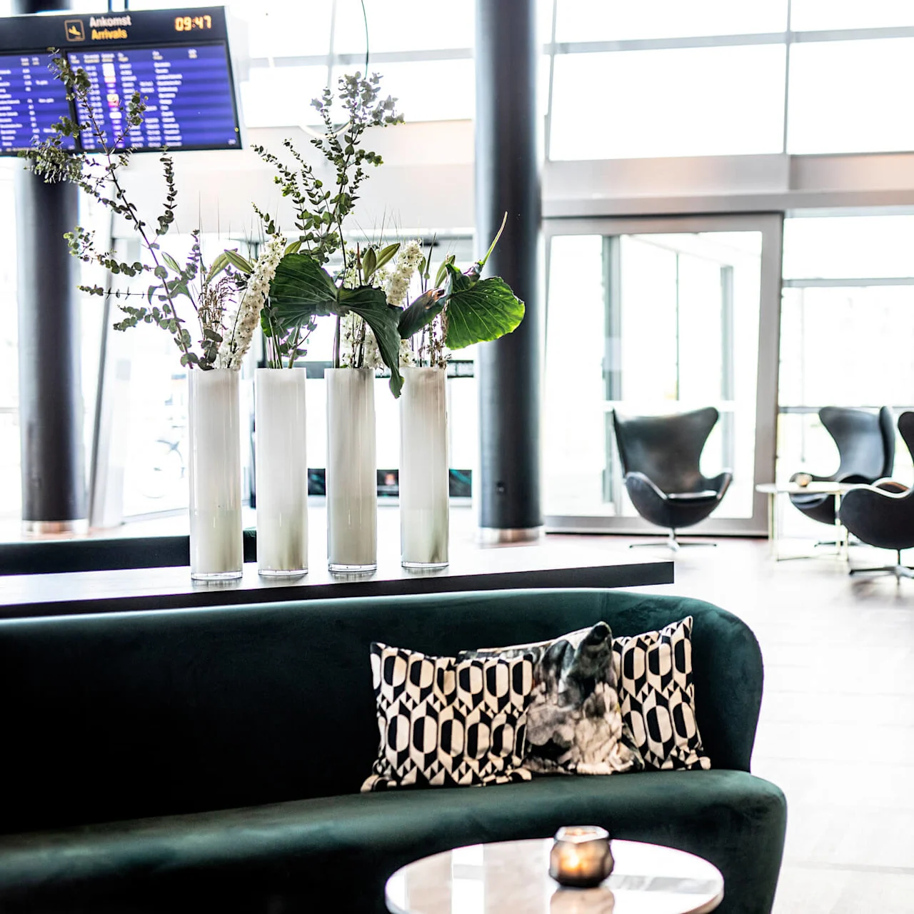 Fyra höga vaser håller frodig grönska på ett bord, omgivet av moderna sittplatser och stora fönster. Den digitala skylten visar "Ankomster Arrivals 09:47".