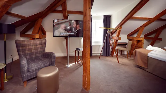 A cozy attic room with exposed wooden beams, a plaid chair, a bed, and a flat-screen TV showing a program with Swedish subtitles: "-Men du hade tvekligt i alla fall? -Ja då."