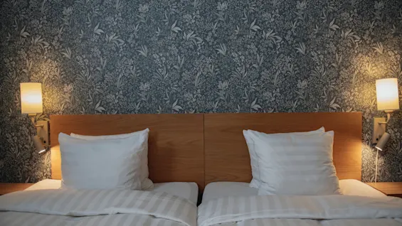 Two neatly made single beds with white pillows and sheets rest against a floral patterned wallpapered wall, illuminated by wall-mounted lamps on either side, creating a cozy bedroom setting.