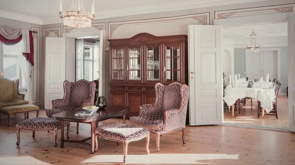 Antikk treskap står elegant, viser glass, ved siden av mønstrede stoler og et sentralt bord i en raffinert stue. Åpne dører leder til en spisestue klar for et måltid.