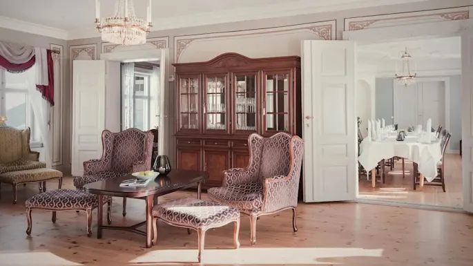 Antique wooden cabinet stands elegantly, displaying glassware, alongside patterned chairs and a central table in a refined living room. Open doors lead to a dining area set for a meal.