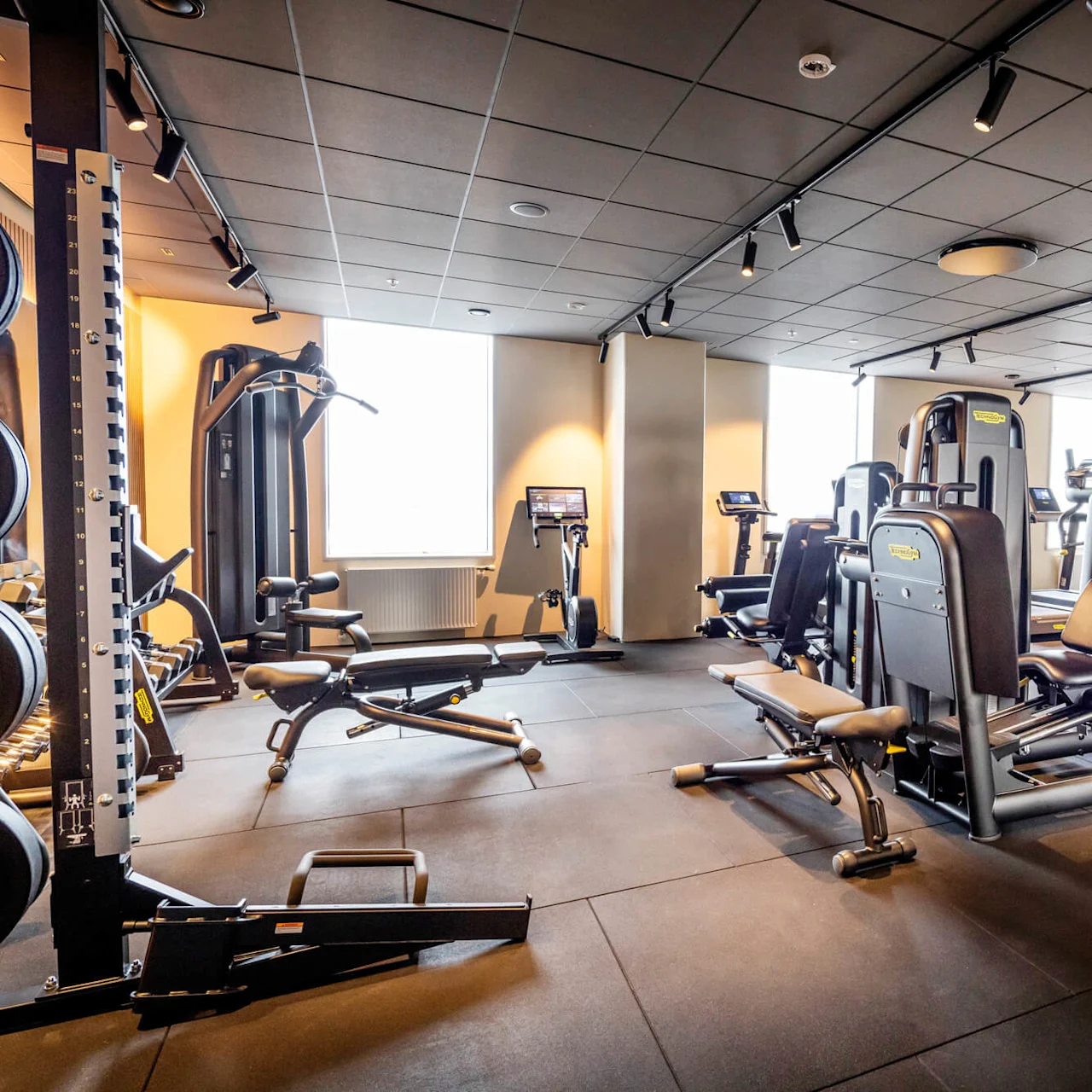 Ett modernt gym med träningsmaskiner och styrketräningsutrustning i ett rymligt, välbelyst rum med svart tak och stora fönster.