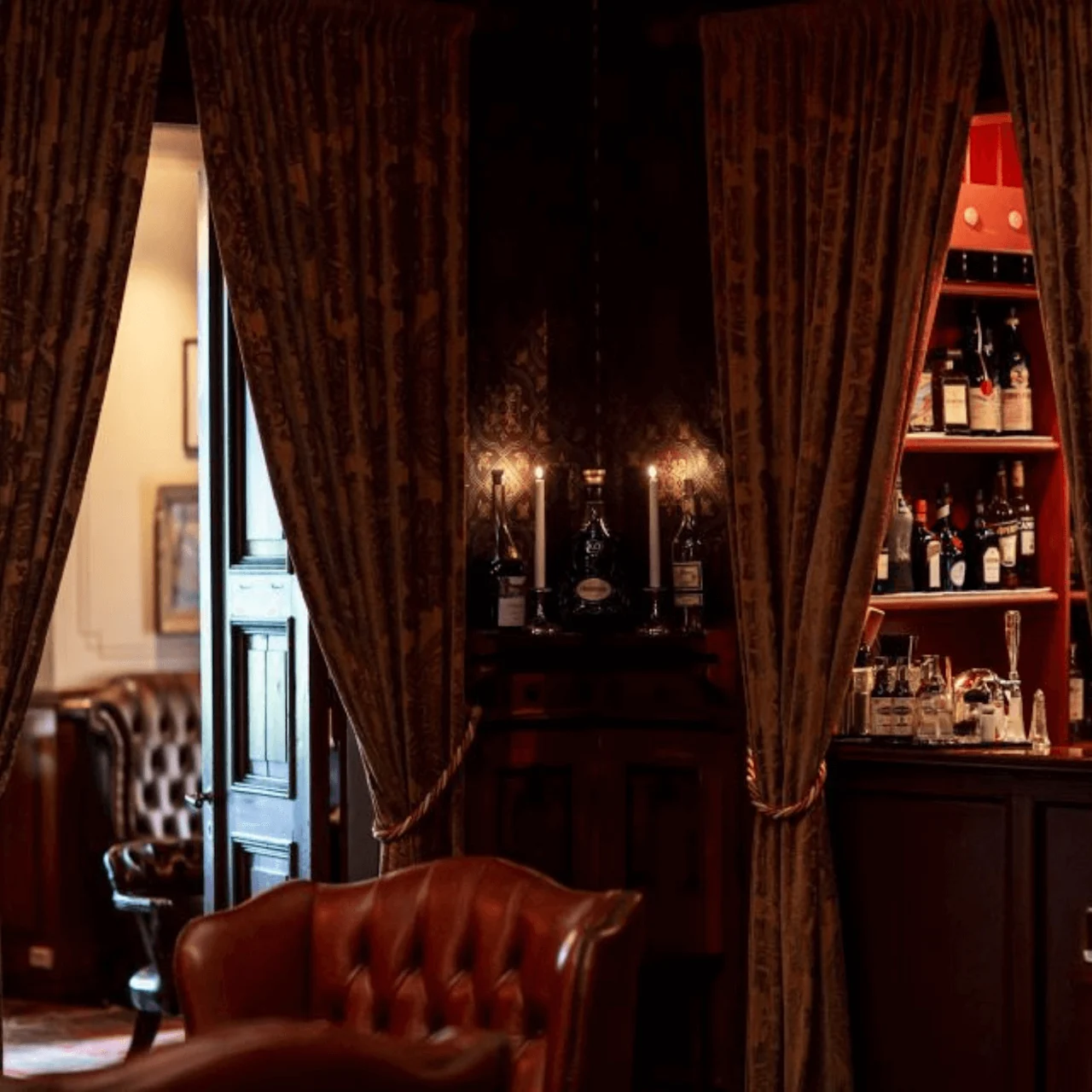 Bar with bottles arranged on shelves and counters, surrounded by rich, dark wood and leather furnishings. Elegant curtains frame the scene, creating a cozy, refined atmosphere.