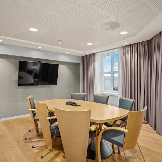 Et konferansebord av tre er omringet av stoler i et møterom, med en stor TV på veggen og vinduer med gardiner som gir naturlig lys.