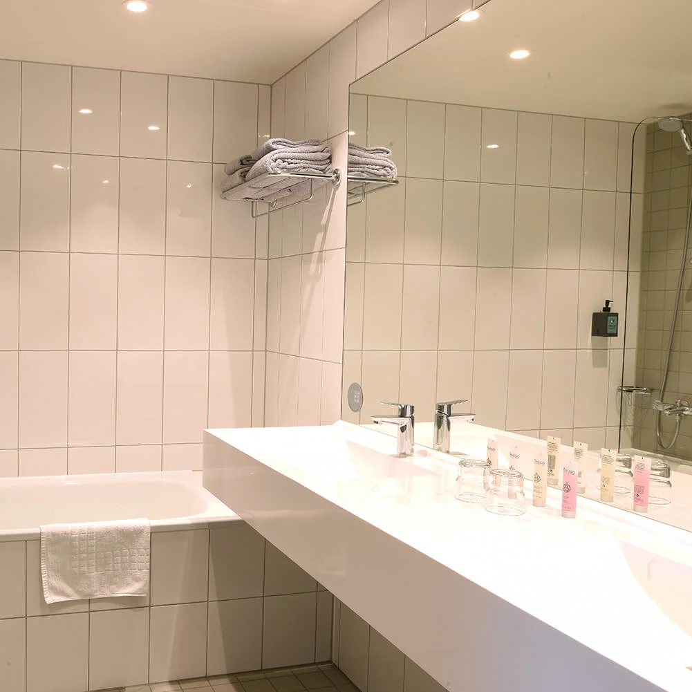 Et hvidt flisebelagt badeværelse har et badekar med en vægmonteret bruser, et stort spejl over en moderne håndvask med toiletartikler, og håndklæder stabler på en hylde.