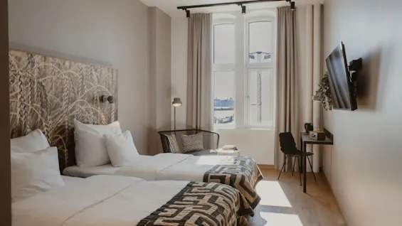 Valoisa hotellihuone, jossa kaksi erillistä sänkyä, kuvioitu sängynpääty, ikkuna näköalalla ja pieni työpöytäalue.
