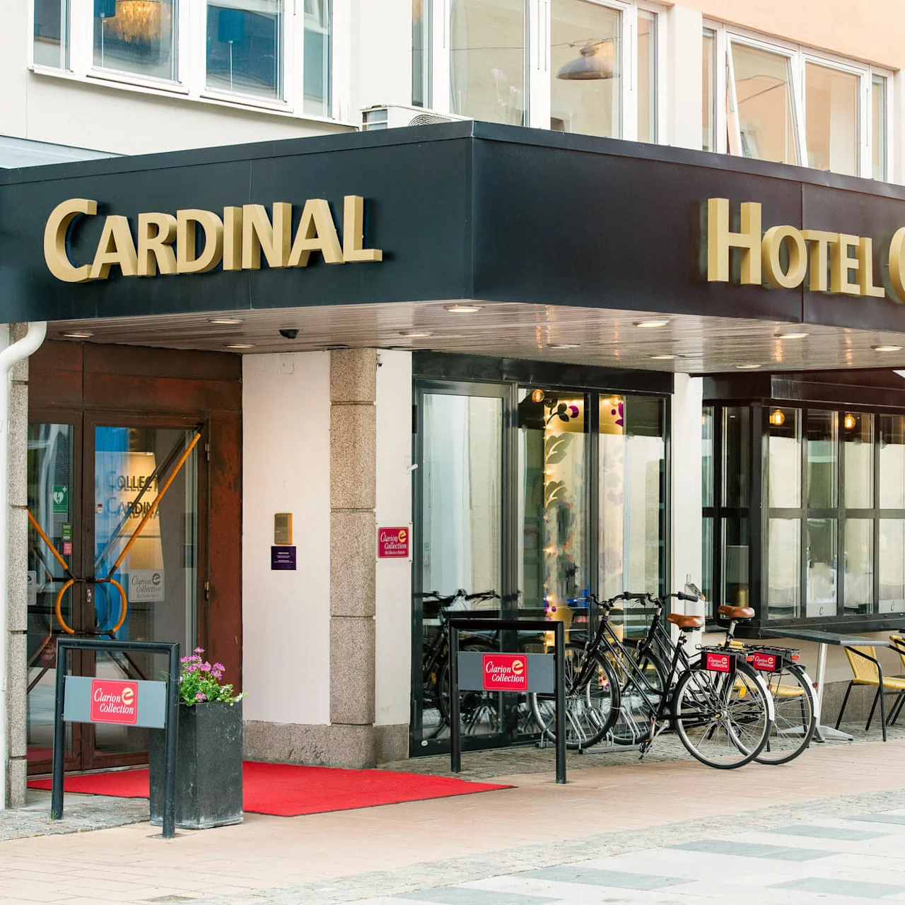 Hotellin sisäänkäynti, jossa on musta ja kultainen kyltti "Cardinal Hotel Cardinal." Polkupyöriä on pysäköitynä ja tuoleja järjestettynä ulkona jalkakäytävällä.