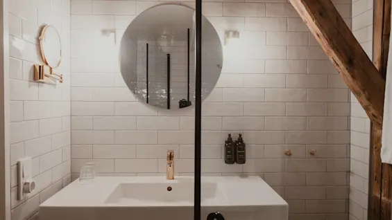 Et moderne badeværelse med hvide fliser, et rundt spejl og en elegant vask med guldfarvede armaturer, med synlige træbjælker.