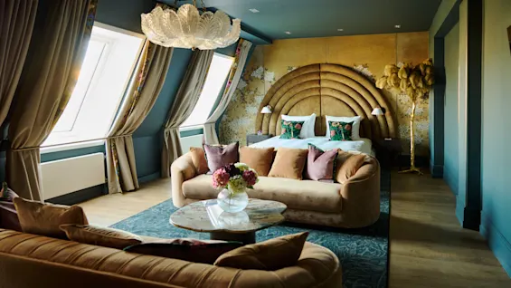 A plush bed with decorative pillows stands against a golden arched headboard. In front, a velvet sofa set surrounds a glass coffee table with flowers. Large windows illuminate the room.