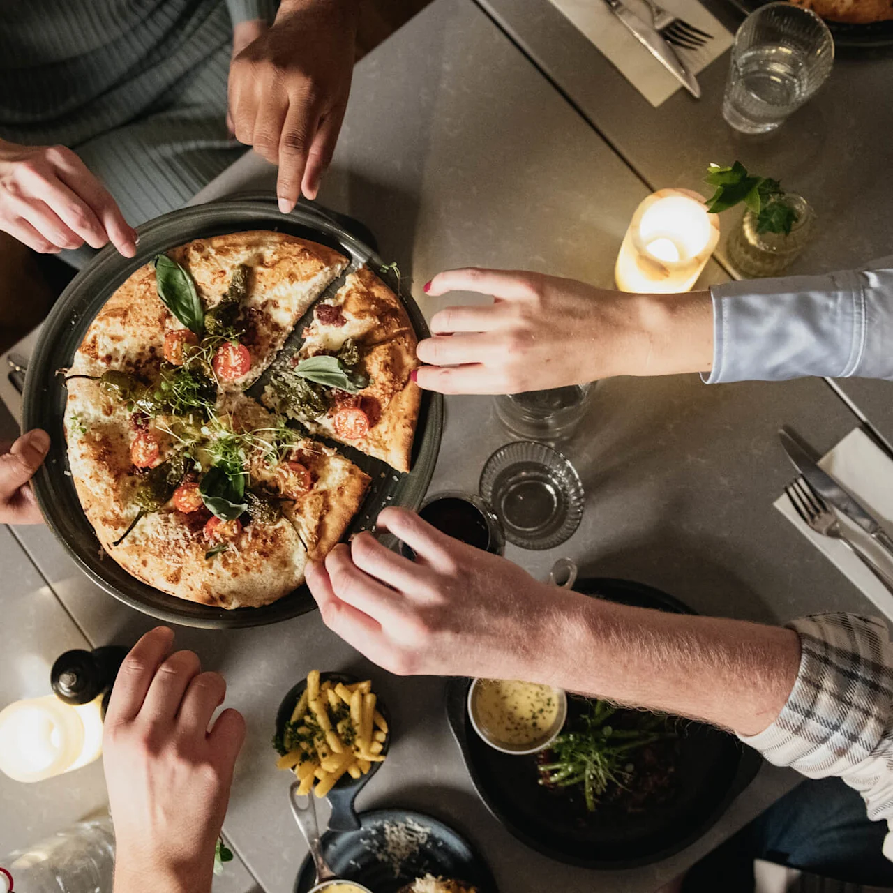Pizza deles av flere hender ved et spisebord. Rundt omkring er det tallerkener og stearinlys, noe som skaper en koselig middagsstemning.