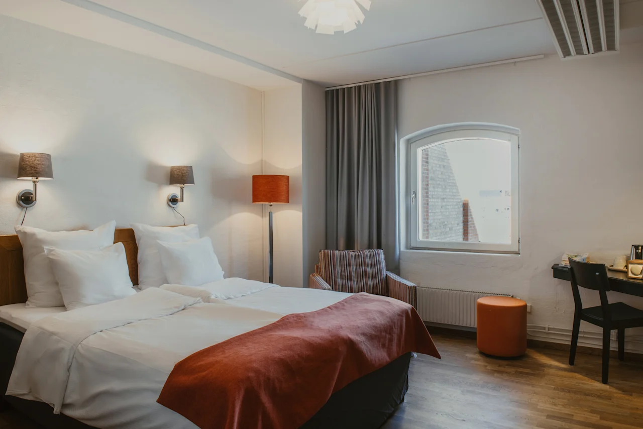 Viihtyisä hotellihuone, jossa on mukava sänky, ikkuna, raidallinen nojatuoli ja pieni työpöytä tuolineen.