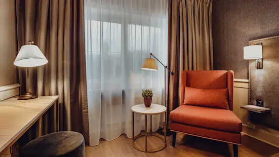 A cozy room with a lit table lamp, sheer curtains, an armchair, side table with a plant, and a wall-mounted lamp.