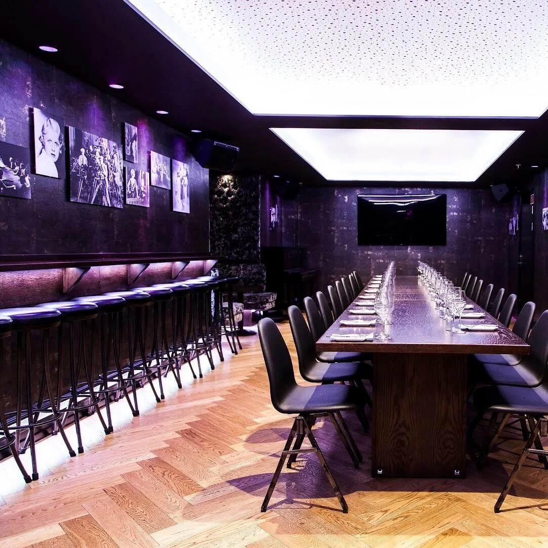 A long, wooden dining table is set with glasses and napkins, surrounded by chairs in a dimly lit room. Black-and-white photos decorate the dark walls, while high stools line the bar.
