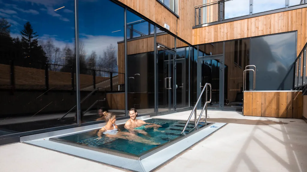 Two people are relaxing in an outdoor hot tub beside a modern wooden building under a clear blue sky at Lily Country Club.